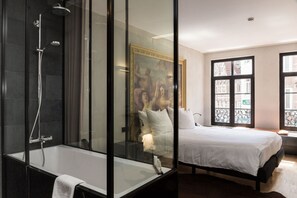 A modern bathroom with a glass-enclosed shower, a large bed with white bedding, and a view of the city through large windows. - HotelO Kathedral Antwerp (Antwerp)