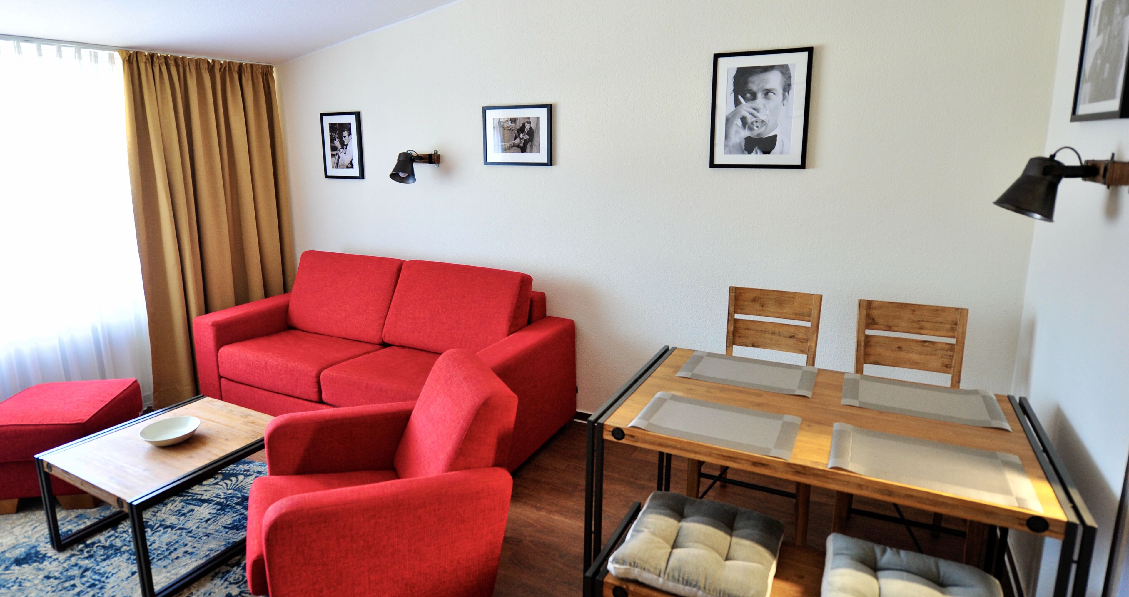 Una sala de estar moderna con un sofá rojo, una mesa de comedor con superficie de vidrio y sillas, y una pequeña mesa de centro. Hay cuadros enmarcados en la pared y una lámpara sobre la mesa.
