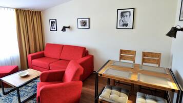Una sala de estar moderna con un sofá rojo, una mesa de comedor con superficie de vidrio y sillas, y una pequeña mesa de centro. Hay cuadros enmarcados en la pared y una lámpara sobre la mesa.