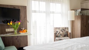 Habitación de hotel con cama, una silla, un televisor montado en la pared, un escritorio con un jarrón de flores y frutas, y una ventana con cortinas traslúcidas.
