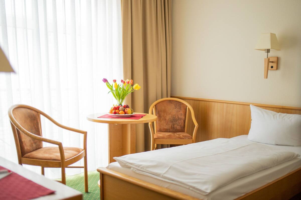 A hotel room with a bed, two chairs, a small table with fruit and flowers, and a lamp on the wall.