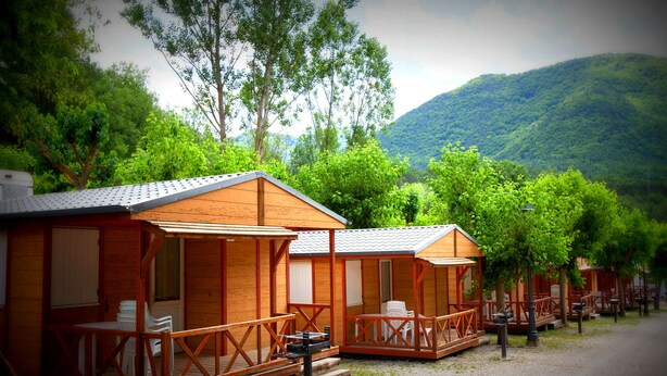 A row of wooden cabins with outdoor seating, set against a backdrop of lush greenery and mountains. - Camping L'Espelt (La Pobla de Lillet)