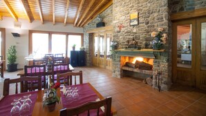 A dining area with wooden tables and chairs, a stone fireplace, and a view of the outside through large windows. - Camping L'Espelt (La Pobla de Lillet)