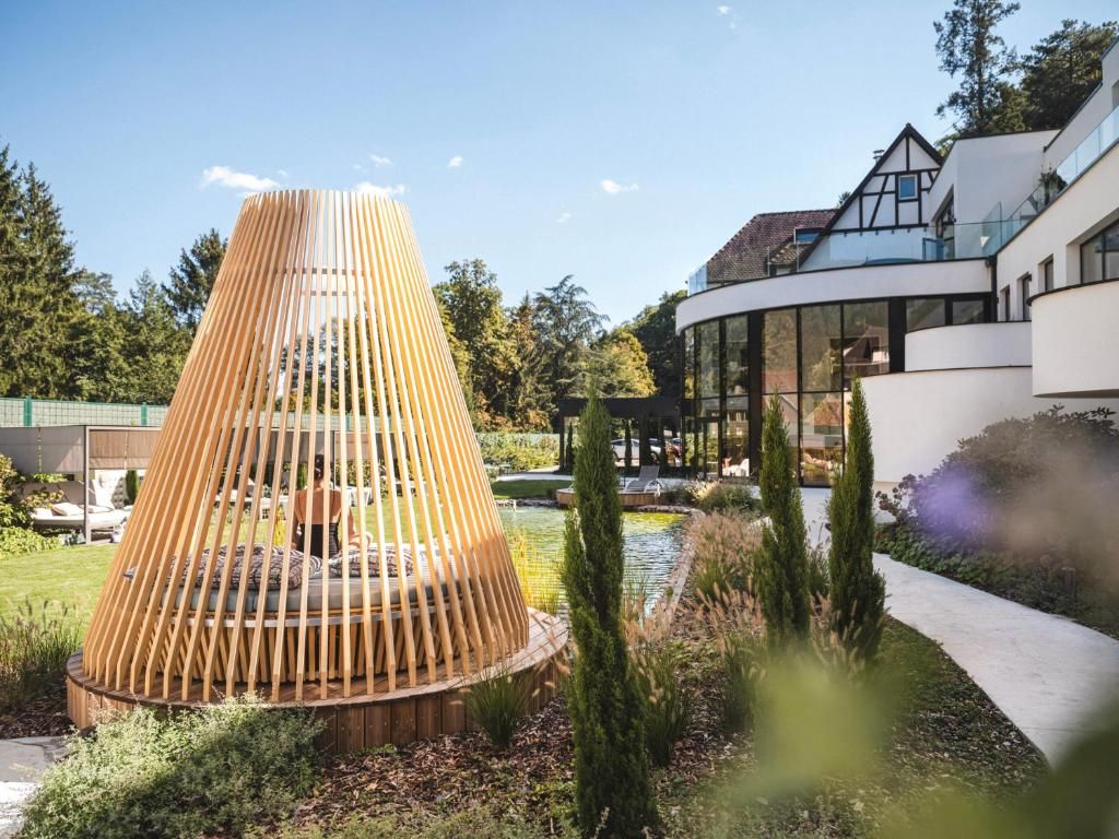 A modern building with a unique conical structure in the foreground, surrounded by a well-maintained garden and a body of water.