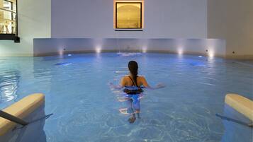 A person in a large indoor pool with clear water, a framed picture on the wall, and a wooden bench on the side.