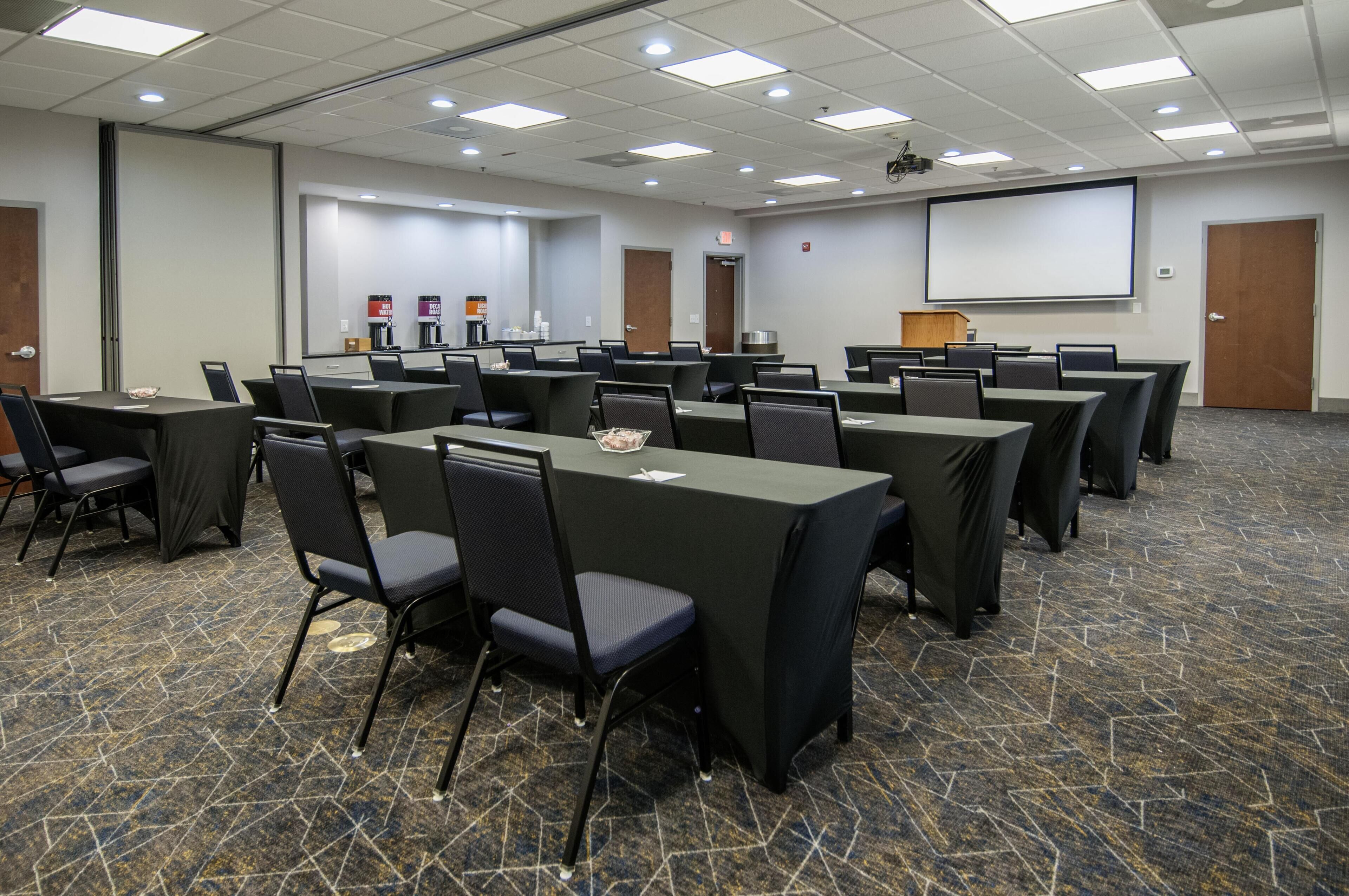 A conference room with multiple tables and chairs, a projector screen, and a carpeted floor.