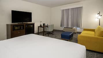 A hotel room with a bed, a television, a desk with a chair, a yellow armchair, and a window with curtains.
