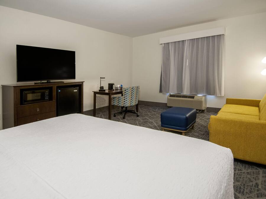 A hotel room with a bed, a television, a desk with a chair, a yellow armchair, and a window with curtains.
