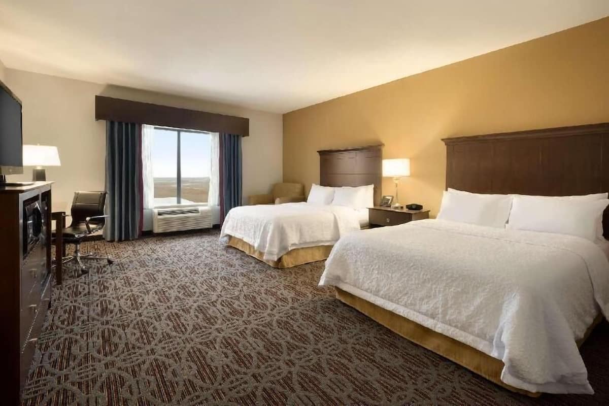 Habitación de hotel con dos camas, un escritorio, una silla, un ventanal con cortinas y vistas al paisaje.