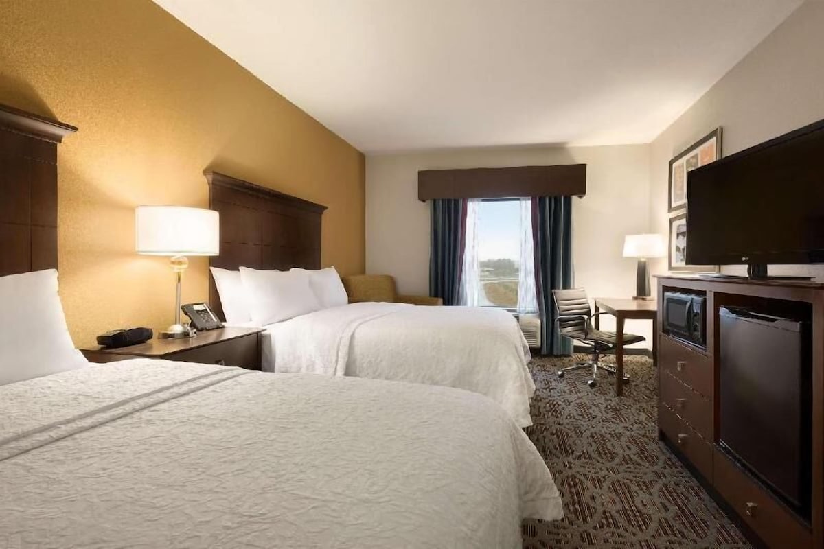 A hotel room with two beds, a desk, a television, and a window with curtains.