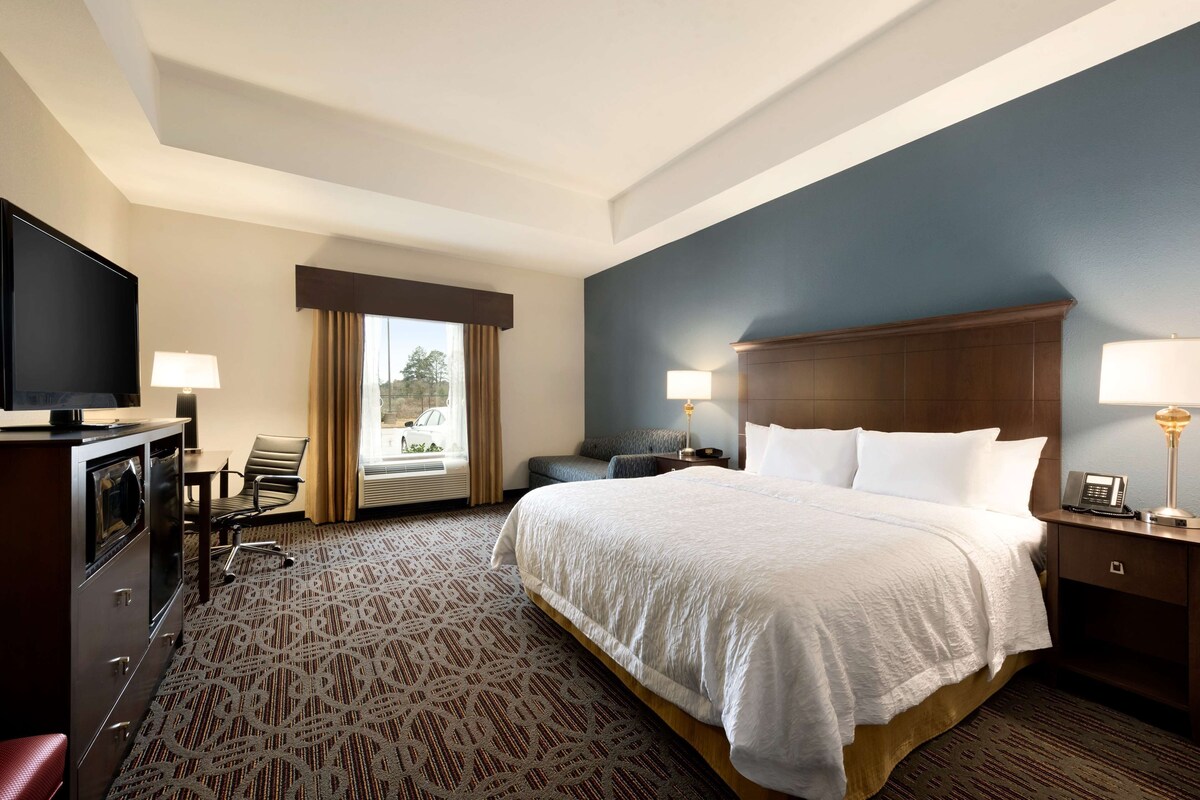 A hotel room with a large bed, a television, a desk with a chair, and a window with curtains.
