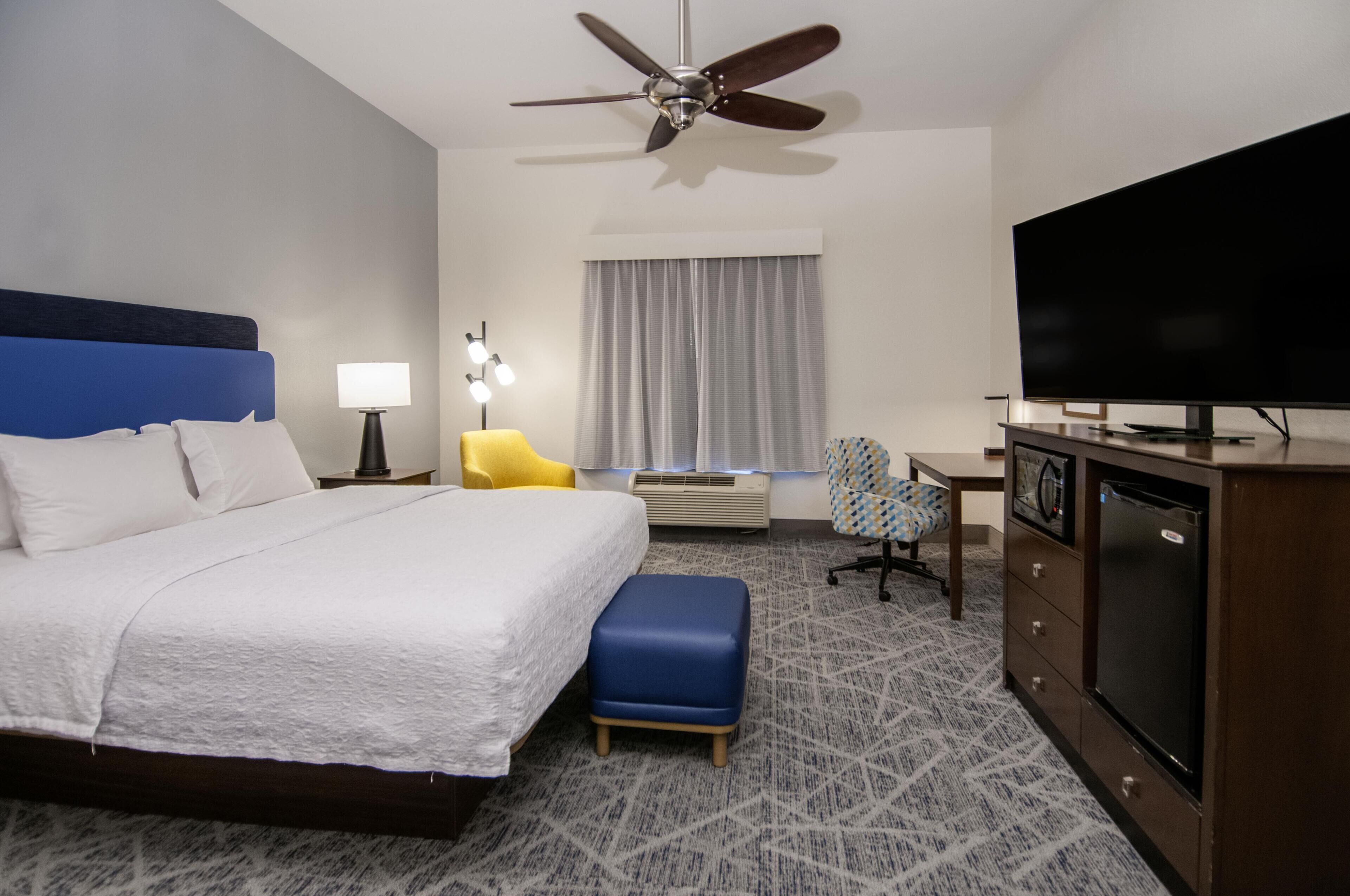 A hotel room with a bed, a desk, a television, a ceiling fan, and an air conditioning unit.