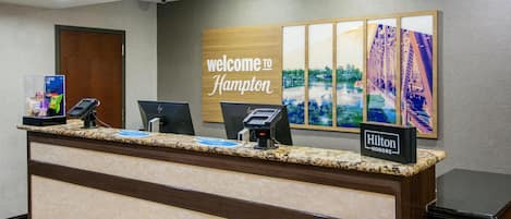 En hotellreception med en skylt som säger ”Välkommen till Hampton”, en digital incheckningsstation och en skylt för ”Hilton Honors”.