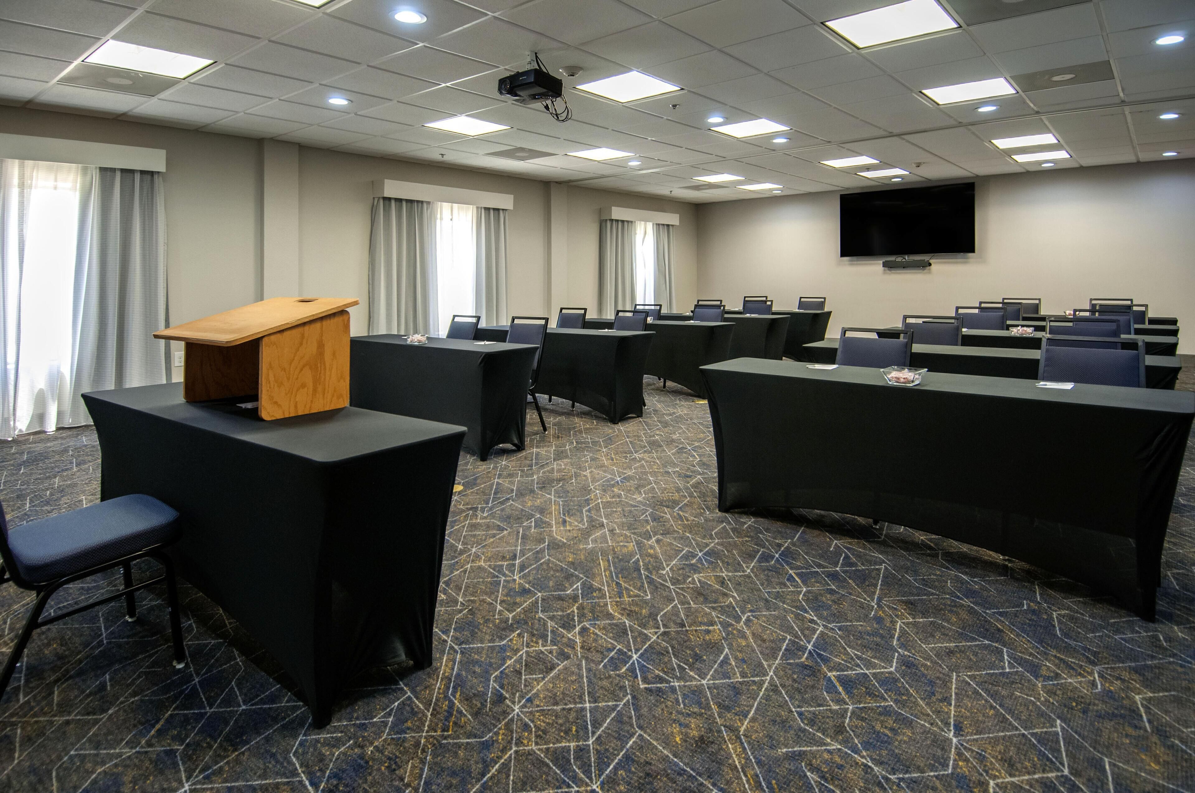 A conference room with multiple tables, chairs, a podium, and a large screen.