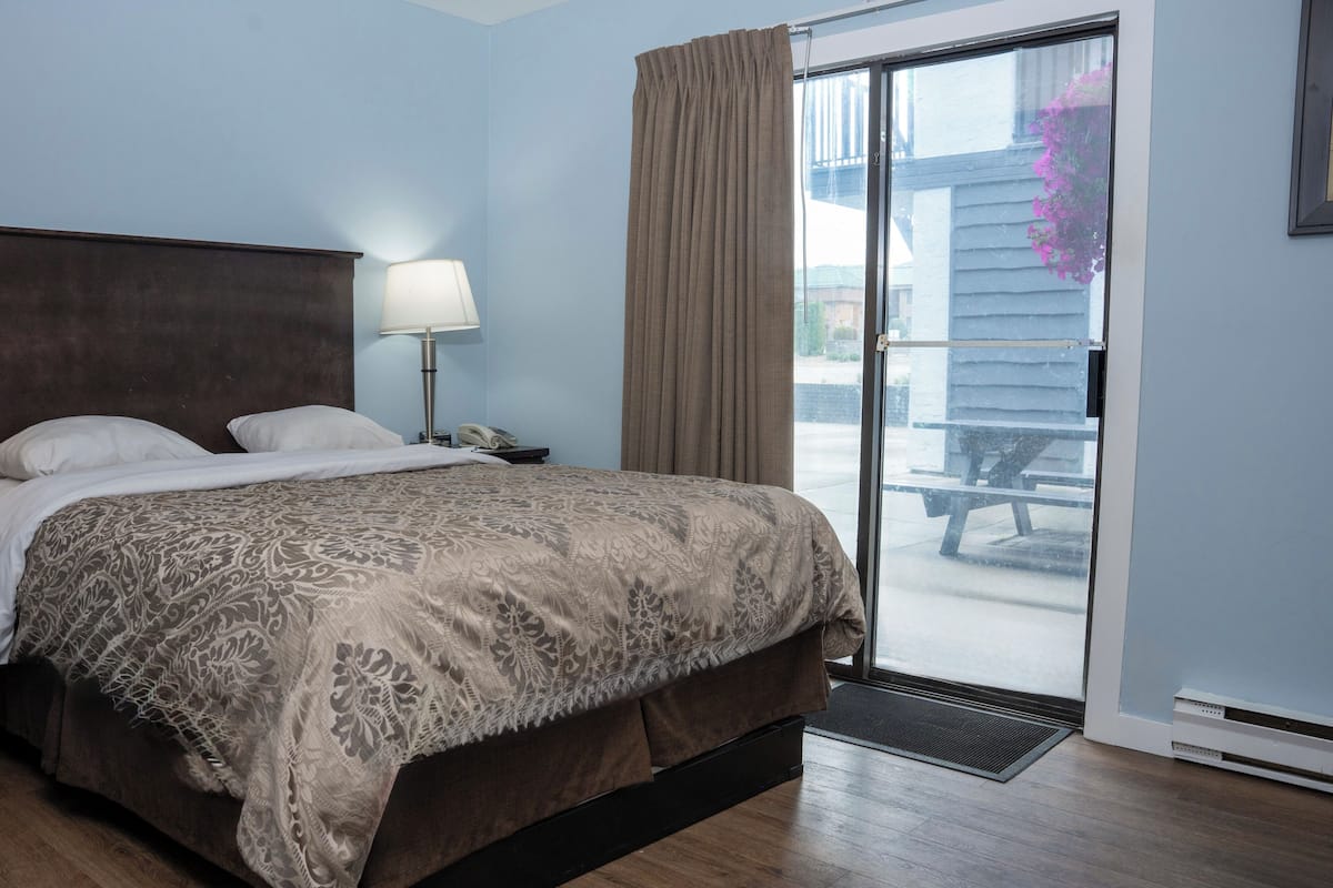A hotel room with a bed, a nightstand, a lamp, a sliding glass door to an outdoor area, and a window with a view of a building and flowers.