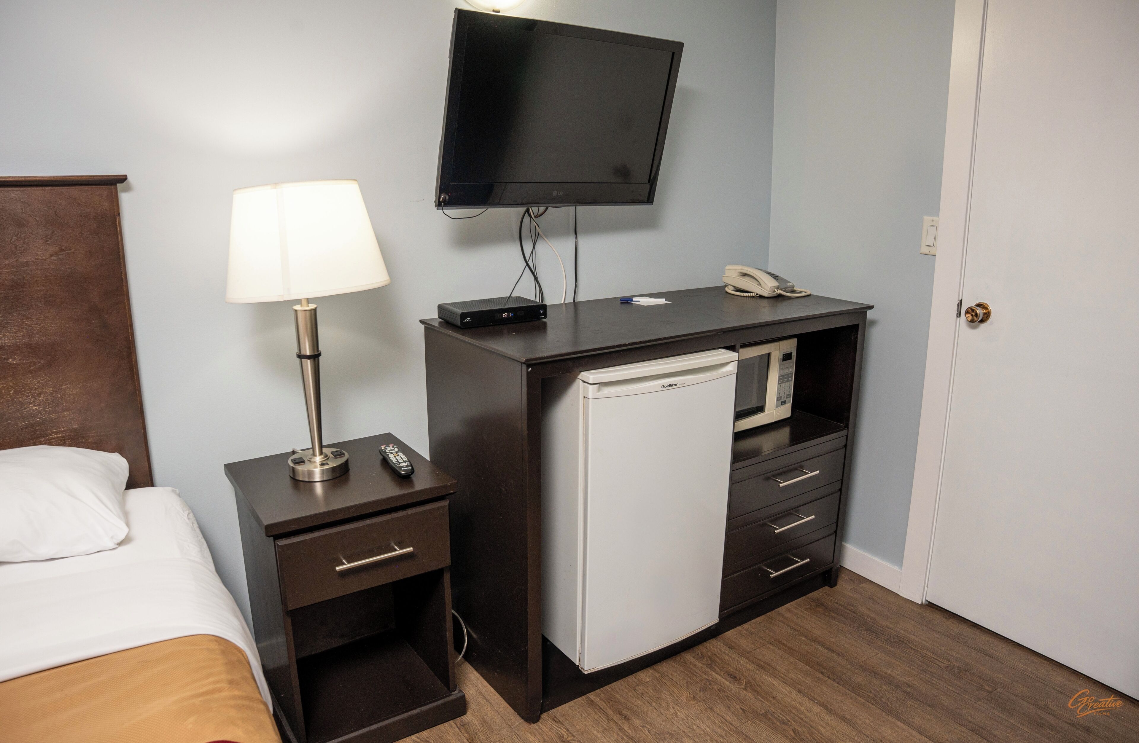 Quarto de hotel com cama, escrivaninha com luminária, televisão, mini-geladeira e telefone.
