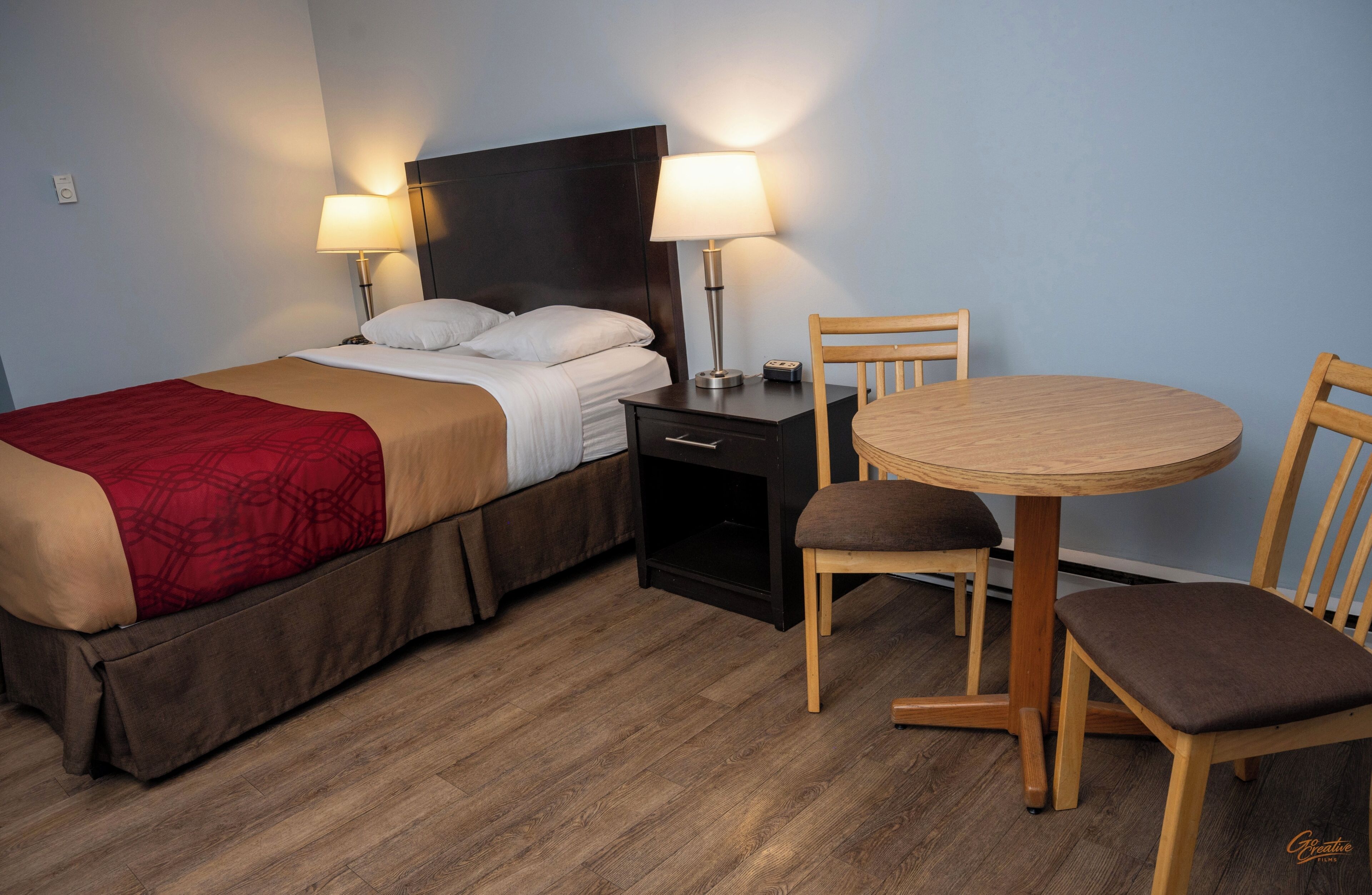 A hotel room with a bed, bedside lamps, a small table with chairs, and a wooden floor.