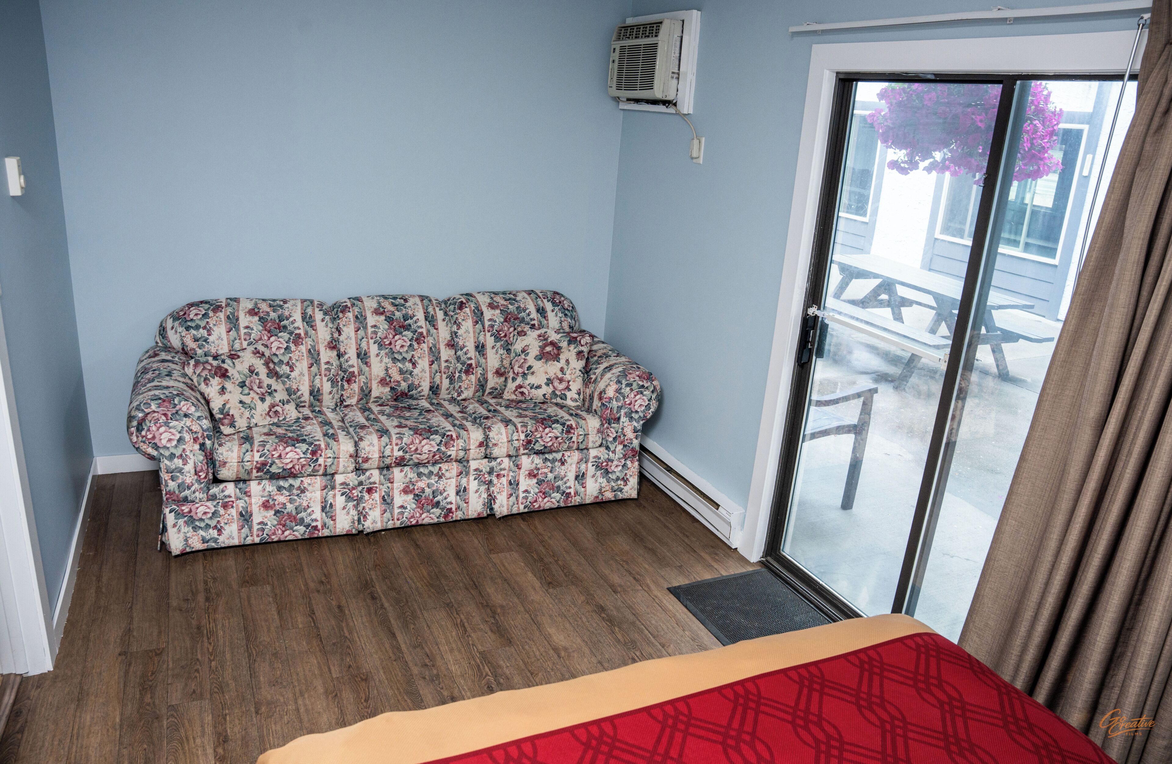 Una habitación con un sofá estampado con motivos florales, piso de madera, puerta corrediza de vidrio que da al exterior y un pequeño equipo de aire acondicionado en la pared.