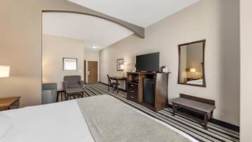 A hotel room with a large bed, a desk with a chair, a small refrigerator, a TV, and a bench.