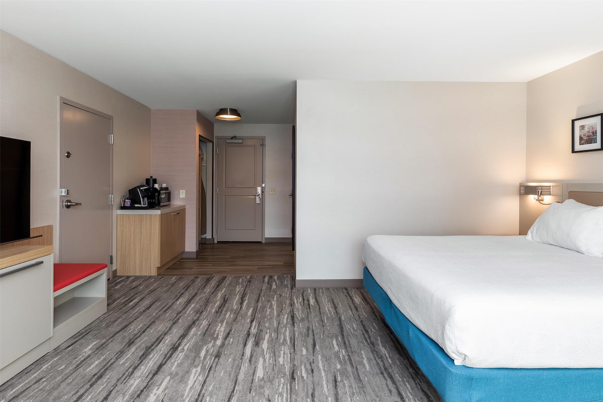 A hotel room with a bed, a TV, a desk with a coffee machine, and a door leading to another room.