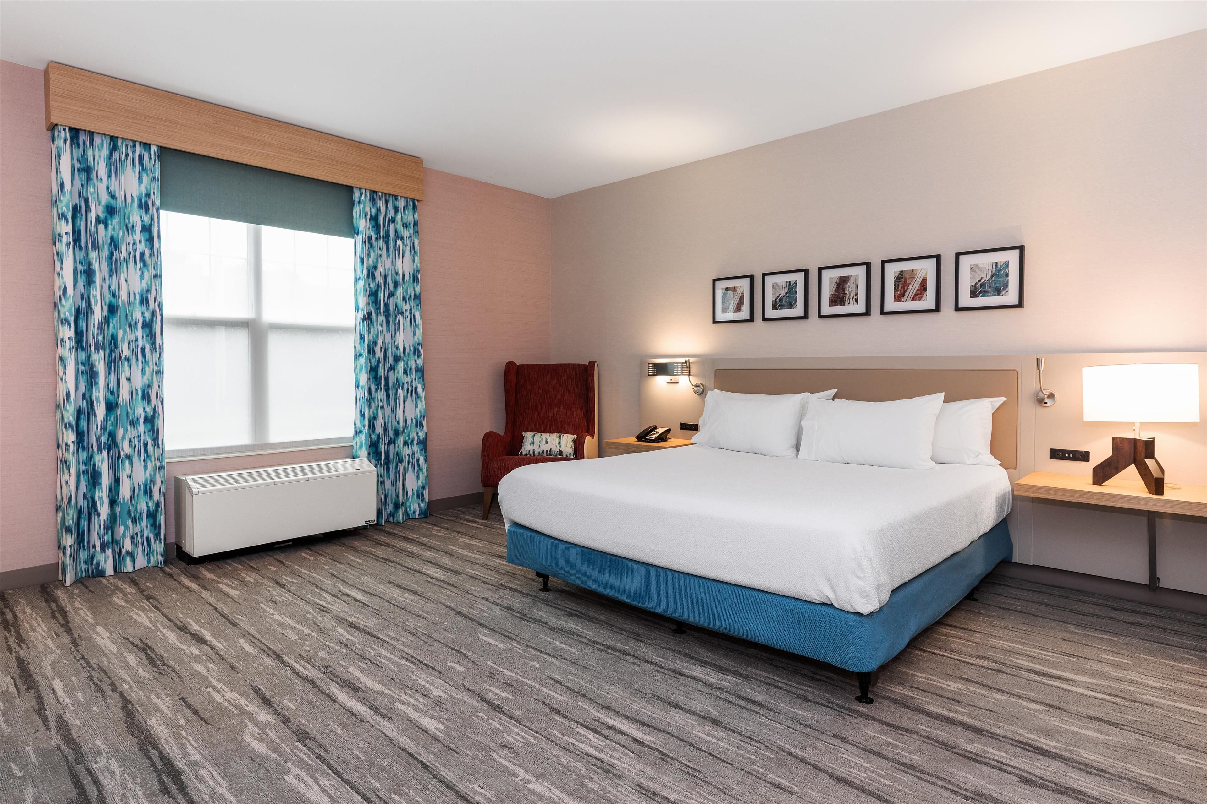 A hotel room with a large bed, a chair, a desk with a lamp, a heater, and a window with curtains.