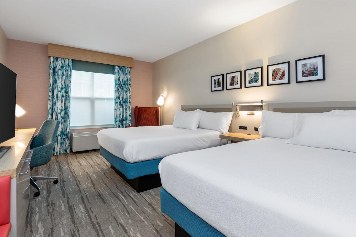 A hotel room with two beds, a desk with a chair, a TV, and a window with curtains.