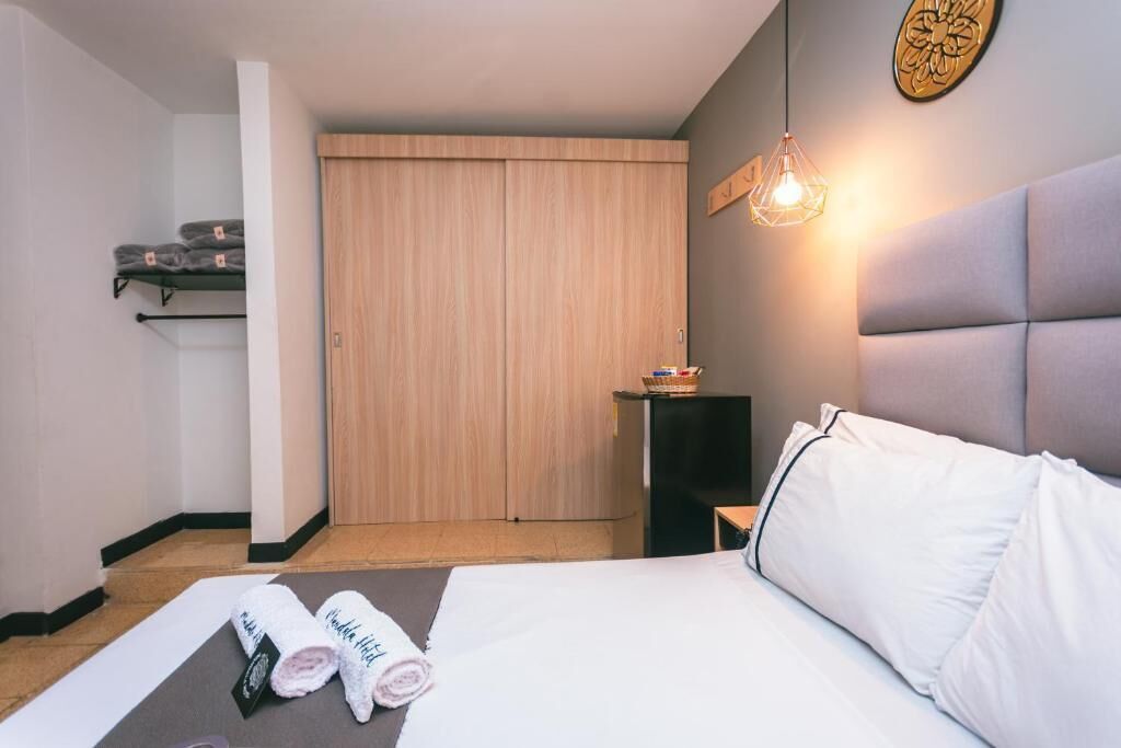 Une chambre d’hôtel comprenant un lit, une table de chevet, un luminaire fixé au mur, une armoire en bois et une étagère avec des serviettes.