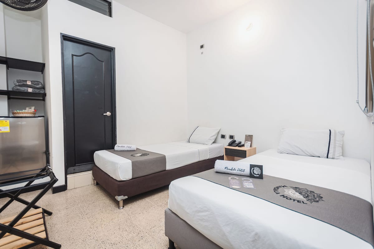 A hotel room with two single beds, a small desk, a refrigerator, and a shelf with items.