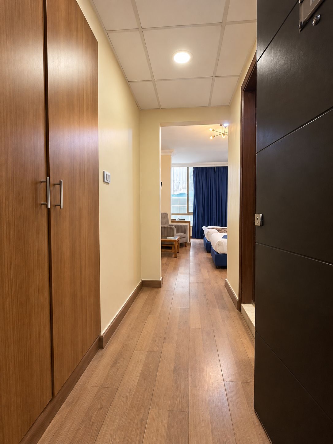 A hotel corridor with wooden flooring, a bed, a desk, and a window with curtains.