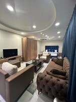 A modern living room with a flat-screen TV, sofa, armchair, and a bed with a headboard.