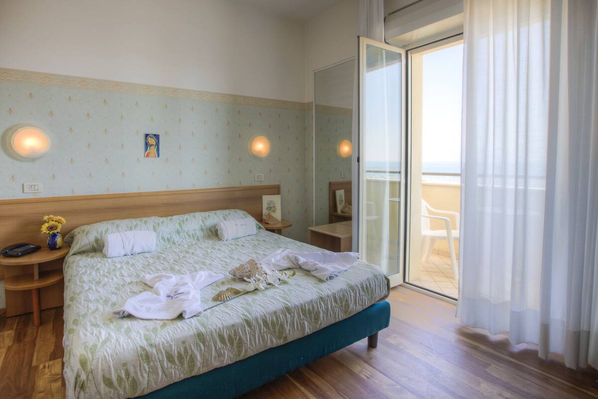 A hotel room with a bed, a nightstand, a small table, and a view of the outside through a glass door.