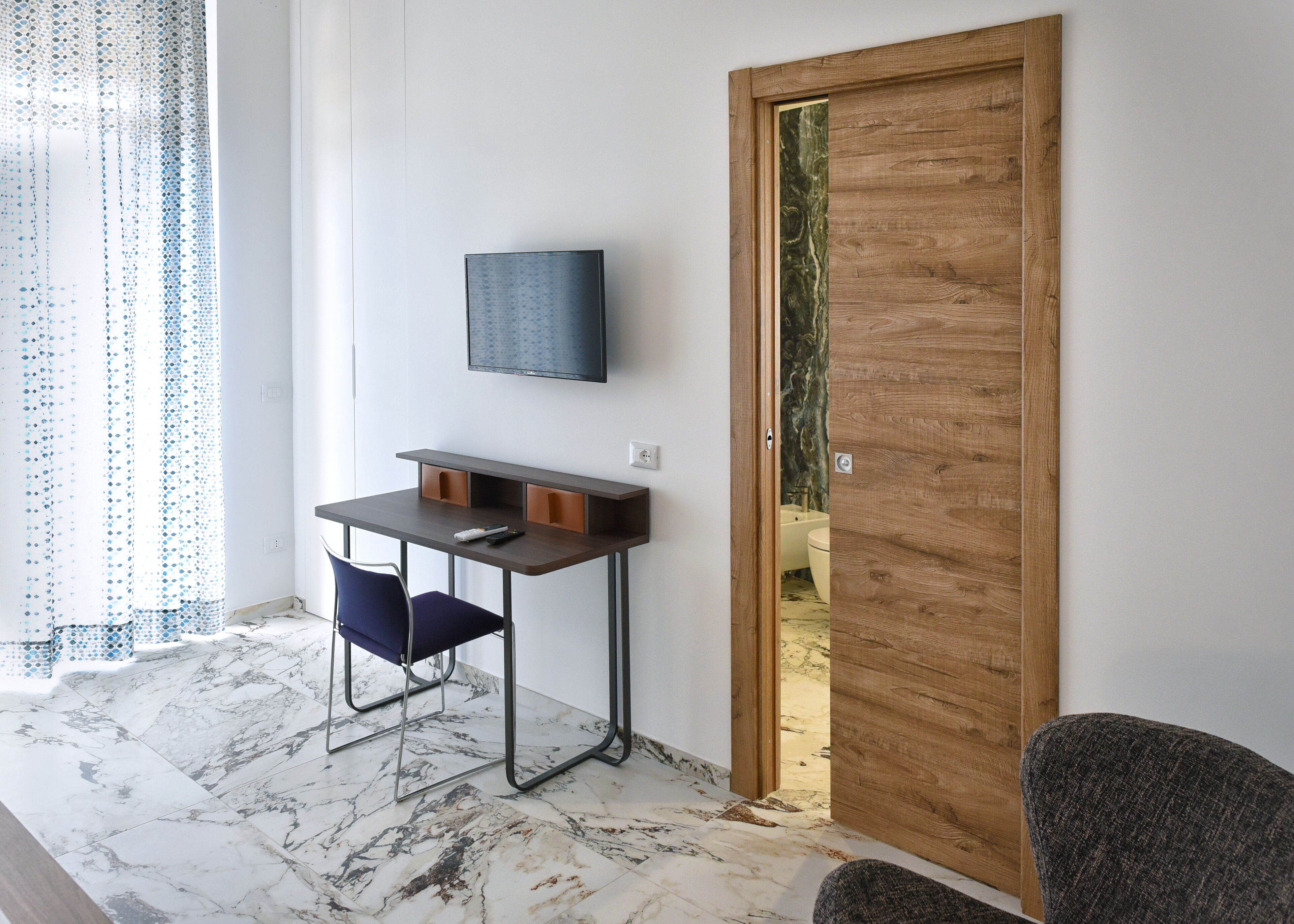A modern room with a wooden door, a TV mounted on the wall, a small desk with a chair, and a bathroom visible through the door.