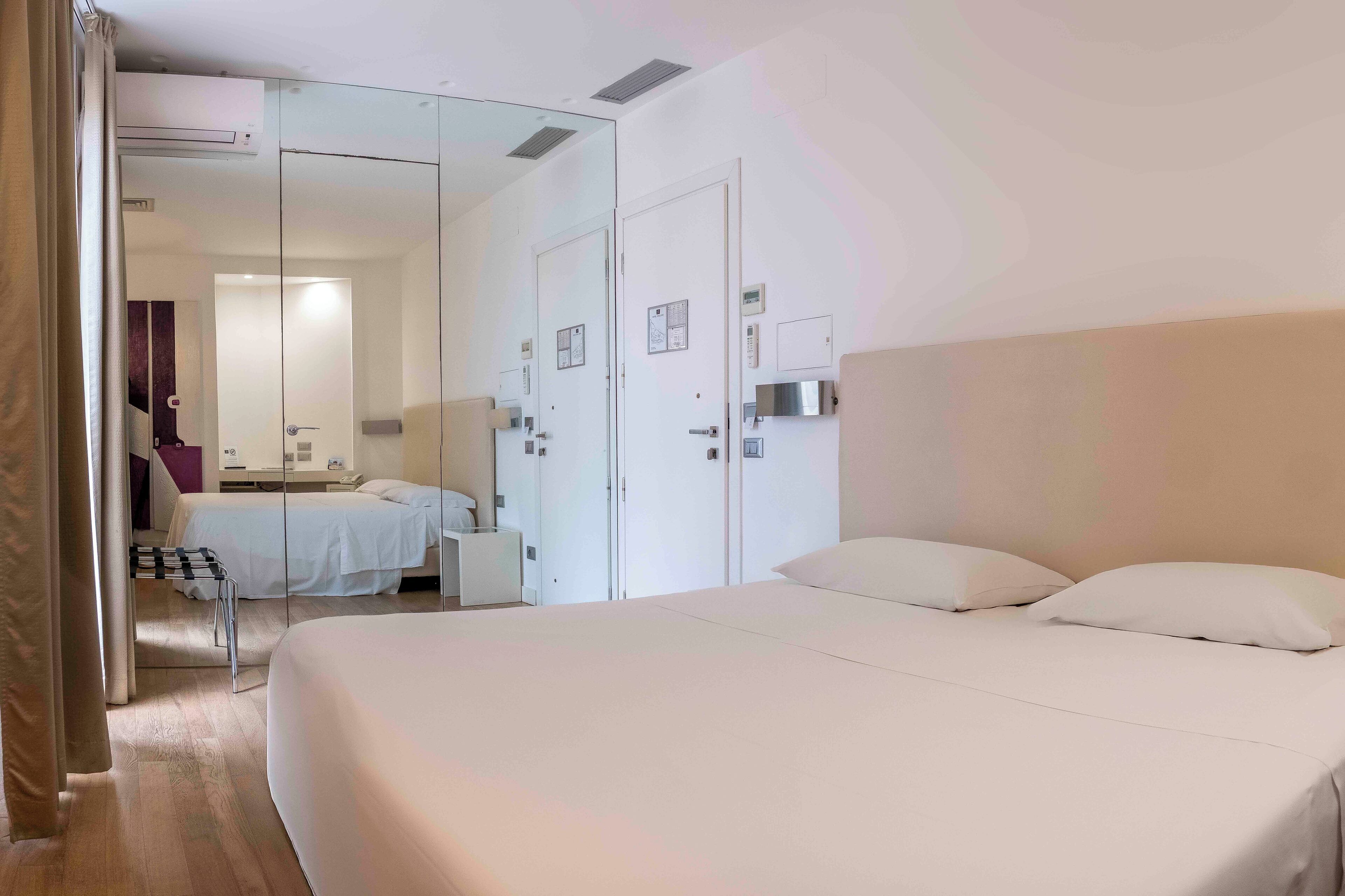 A modern hotel room with a large bed, a small desk, a chair, a mirror, and a wardrobe.