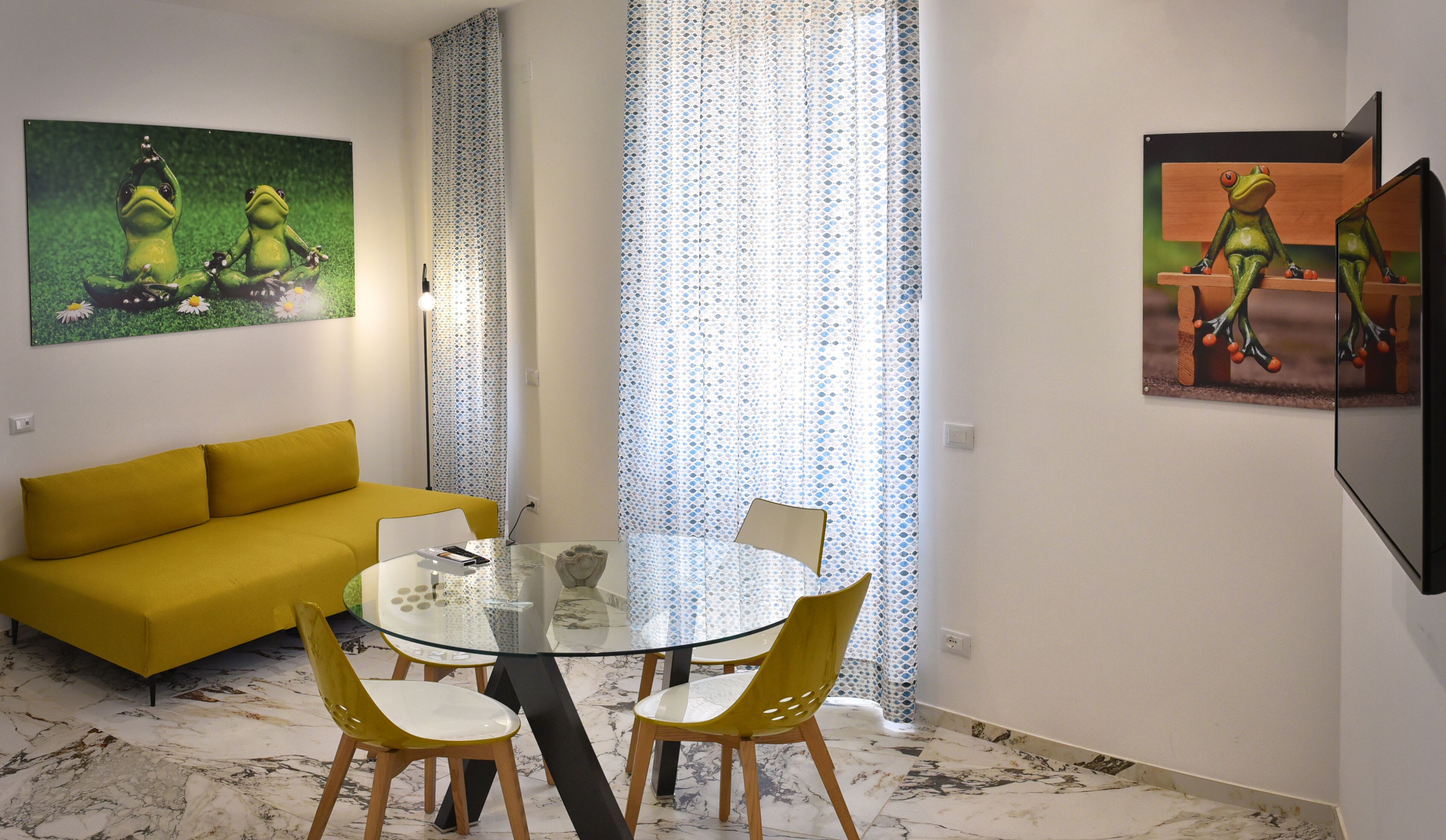 A modern living room with a yellow sofa, a glass dining table with two chairs, and a large window with patterned curtains.