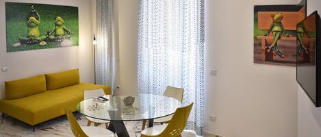 Un salon moderne avec un canapé jaune, une table à manger en verre accompagnée de deux chaises, et une grande fenêtre ornée de rideaux à motifs.