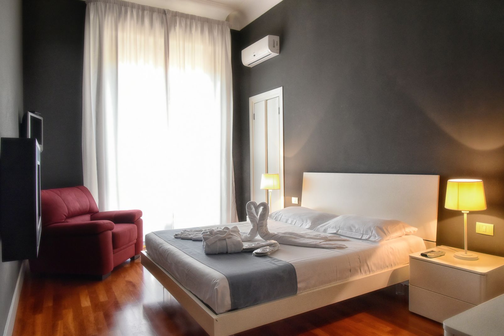Una camera d'albergo con un letto, una poltrona rossa, un comodino con una lampada e un'ampia finestra con tende.