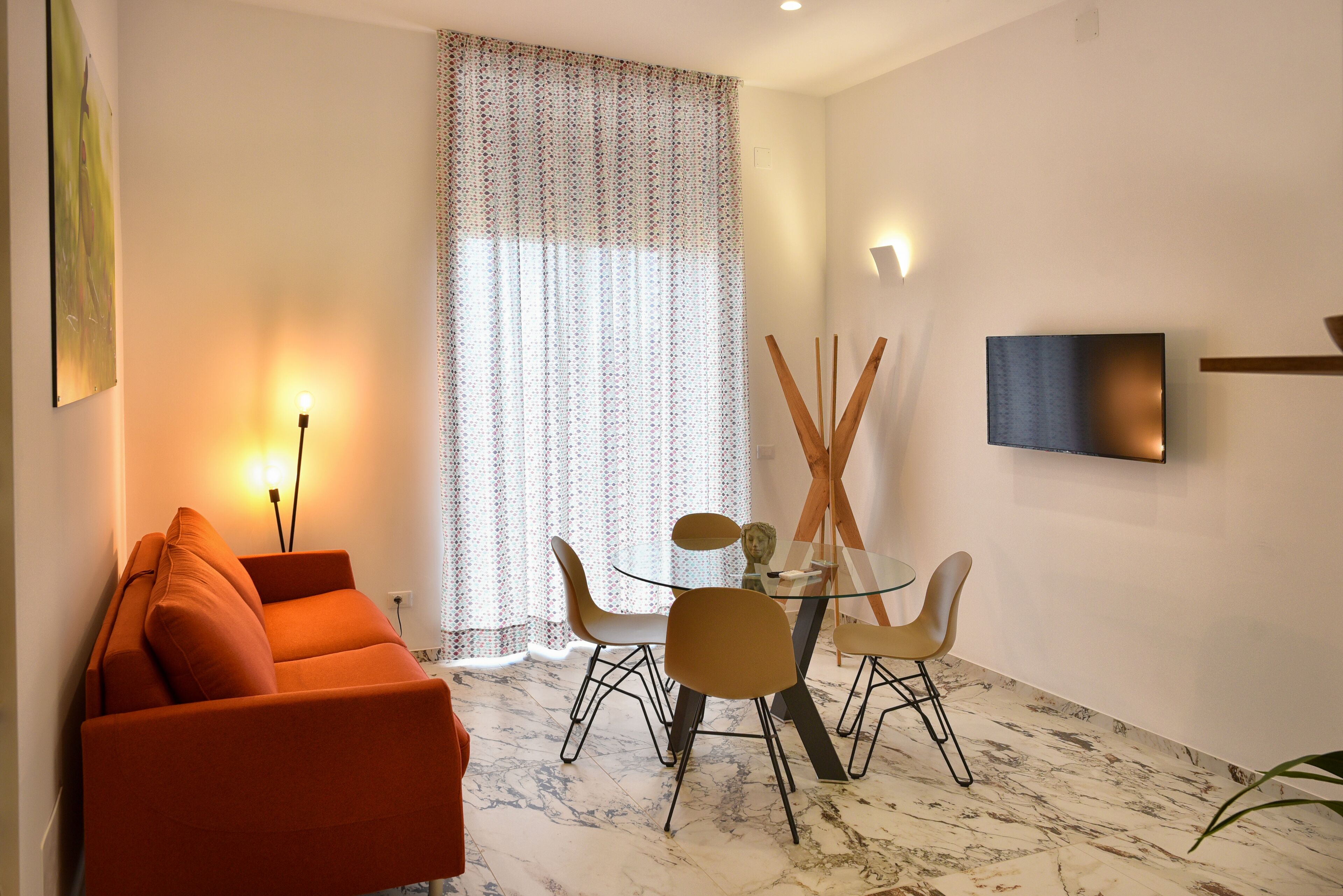 A modern living room with a sofa, a glass dining table, and chairs. There is a large window with curtains, a flat-screen TV mounted on the wall, and a floor lamp.