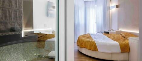 A modern hotel room with a glass-enclosed jacuzzi, a curved bed with a yellow and white bedspread, and a wooden floor.