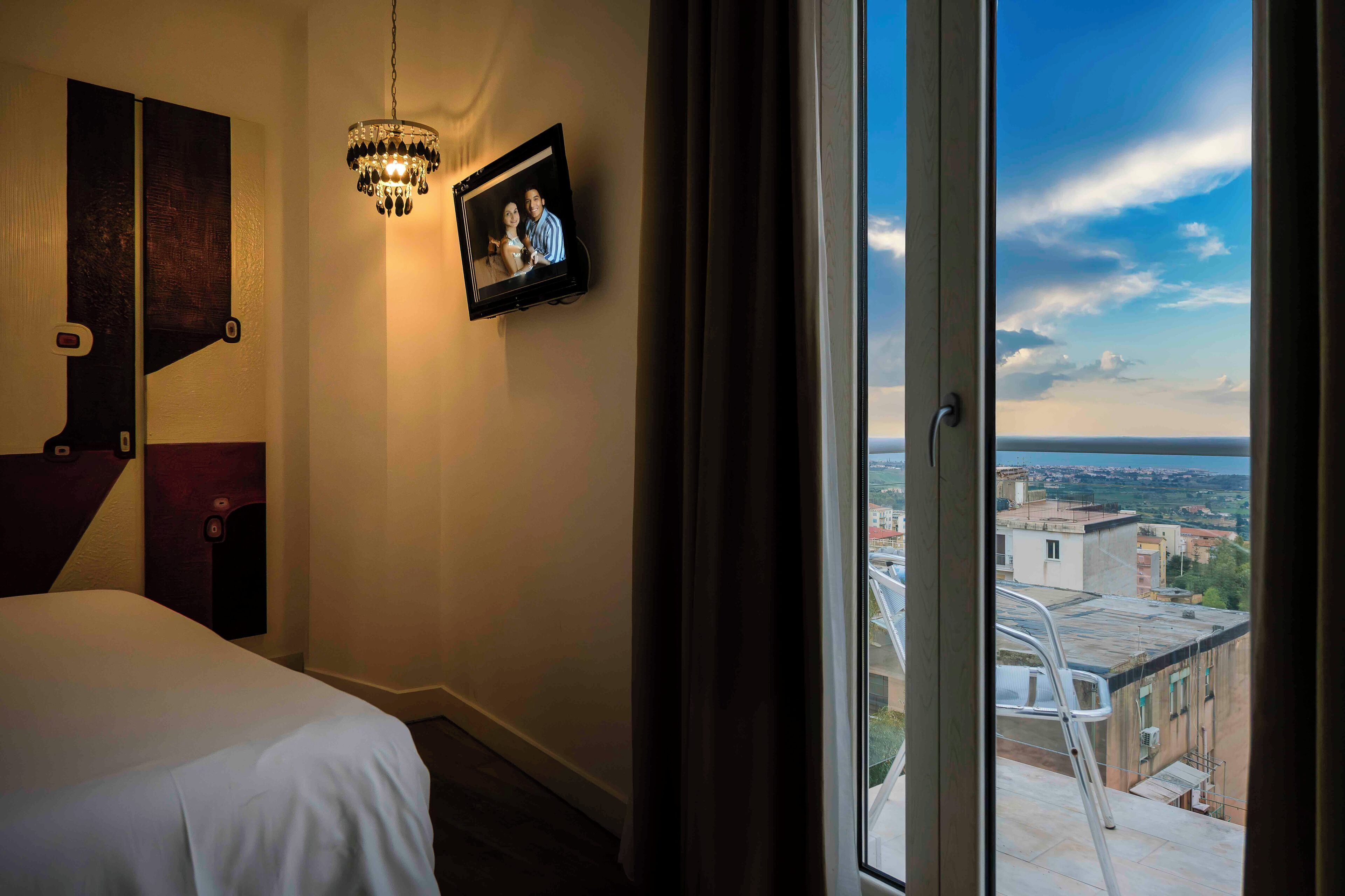 A hotel room with a bed, a mounted television displaying a photo, and a large window offering a city view.