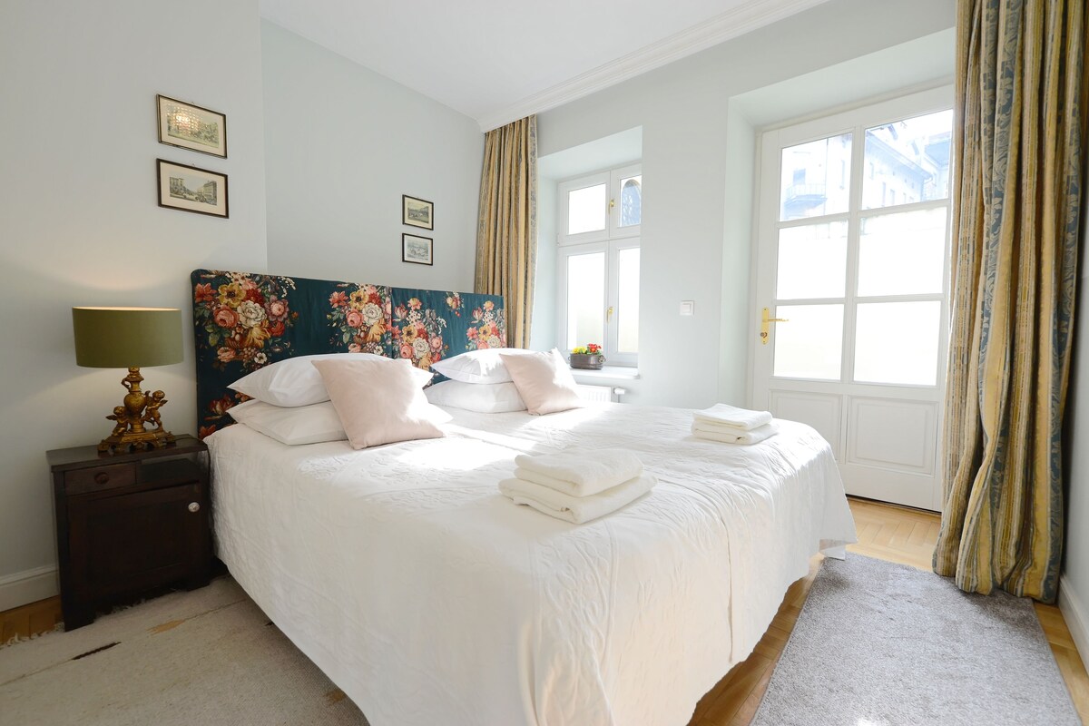 A neatly made bed with white linens and pink pillows, a bedside lamp, and a floral headboard.