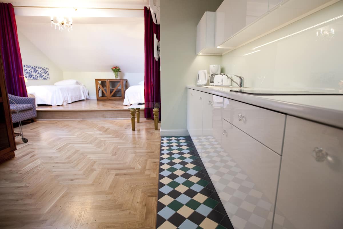 Ein Hotelzimmer mit Holzdielen im Fischgrätmuster, einem Schachbrettmuster aus Fliesen, einem Bett, einem kleinen Tisch mit einer Vase und einer Küchenzeile mit weißen Schränken und einer Arbeitsplatte.