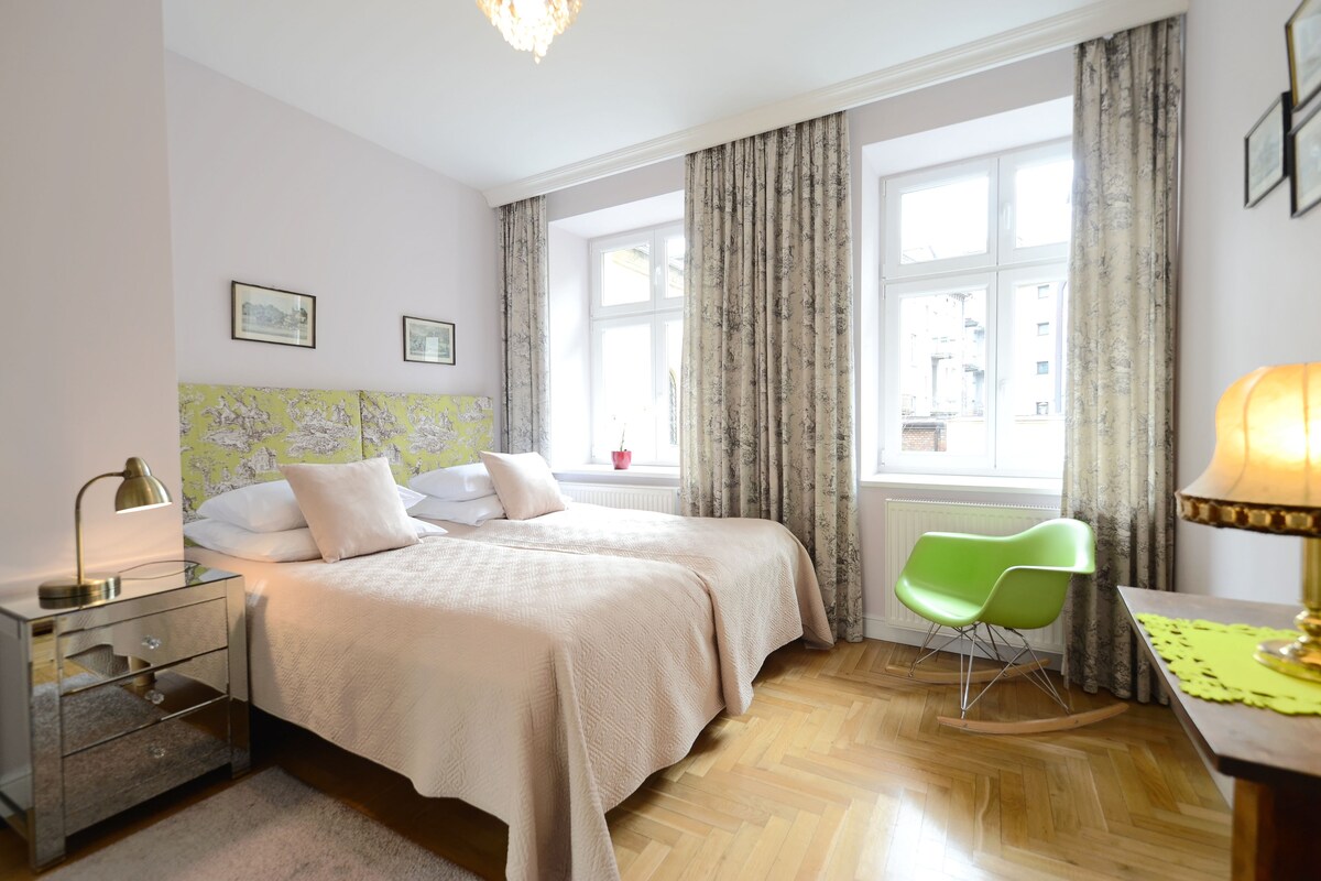 A bedroom with two beds, a green chair, a nightstand, and a window with curtains.