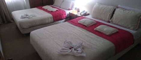 Habitación de hotel con dos camas, cada una con ropa de cama blanca y detalles rosados. Se observa un armario de madera y una mesita de noche con una lámpara y un teléfono.