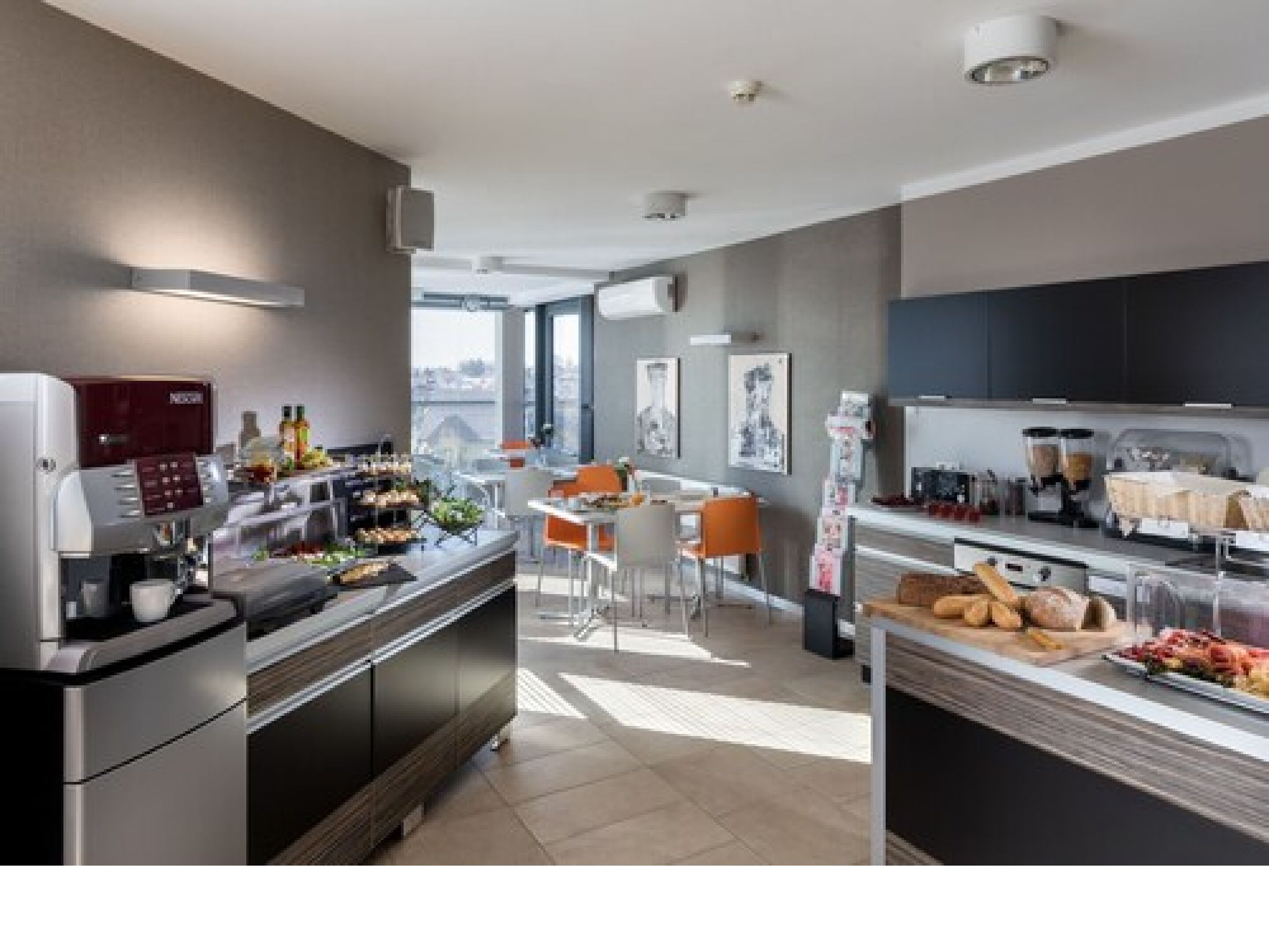 Una cocina moderna con cafetera, un comedor con sillas color naranja y vistas a la ciudad a través de amplios ventanales.