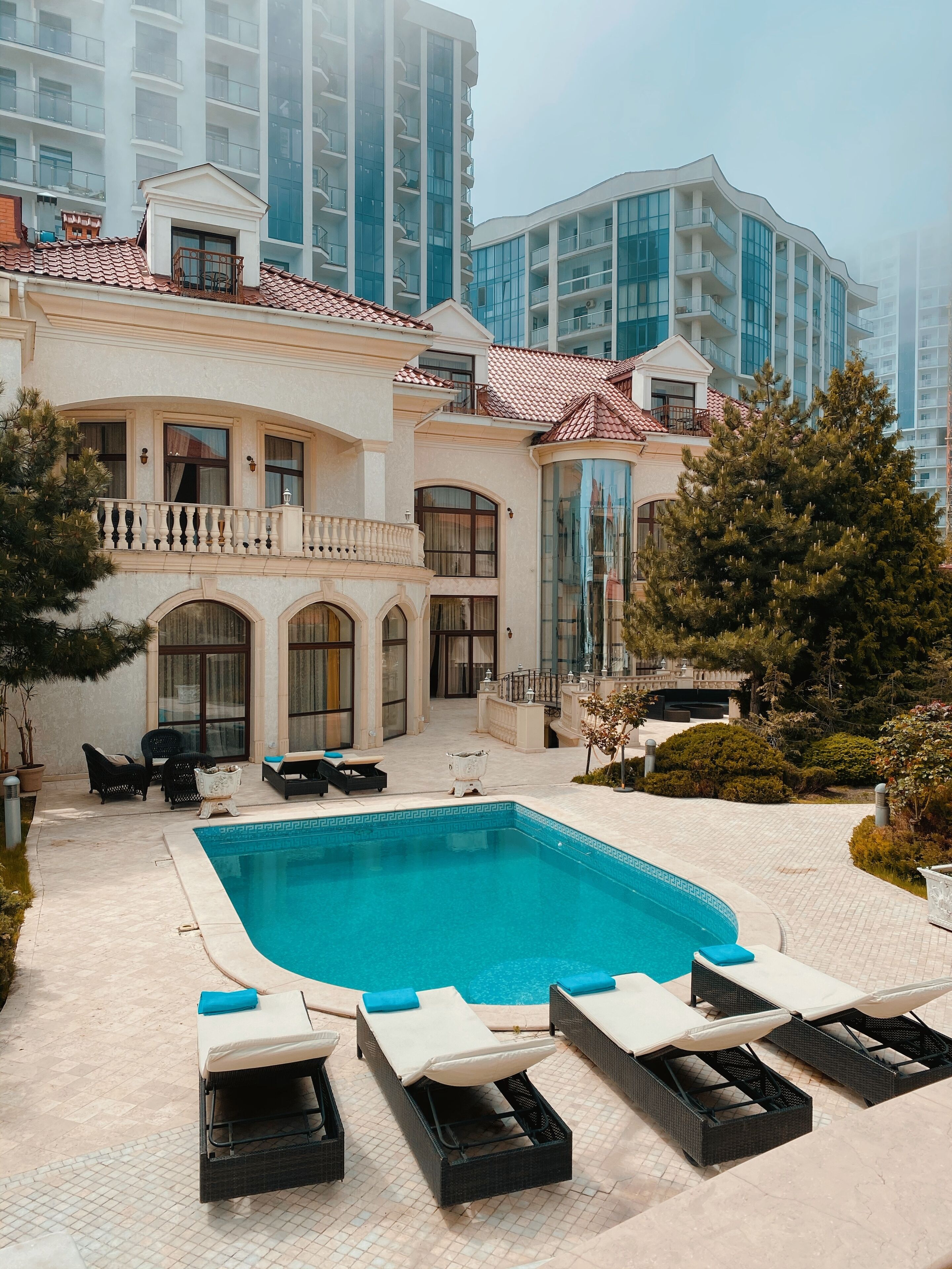 Area piscina con lettini e piscina, circondata da un edificio con balconi e un giardino.