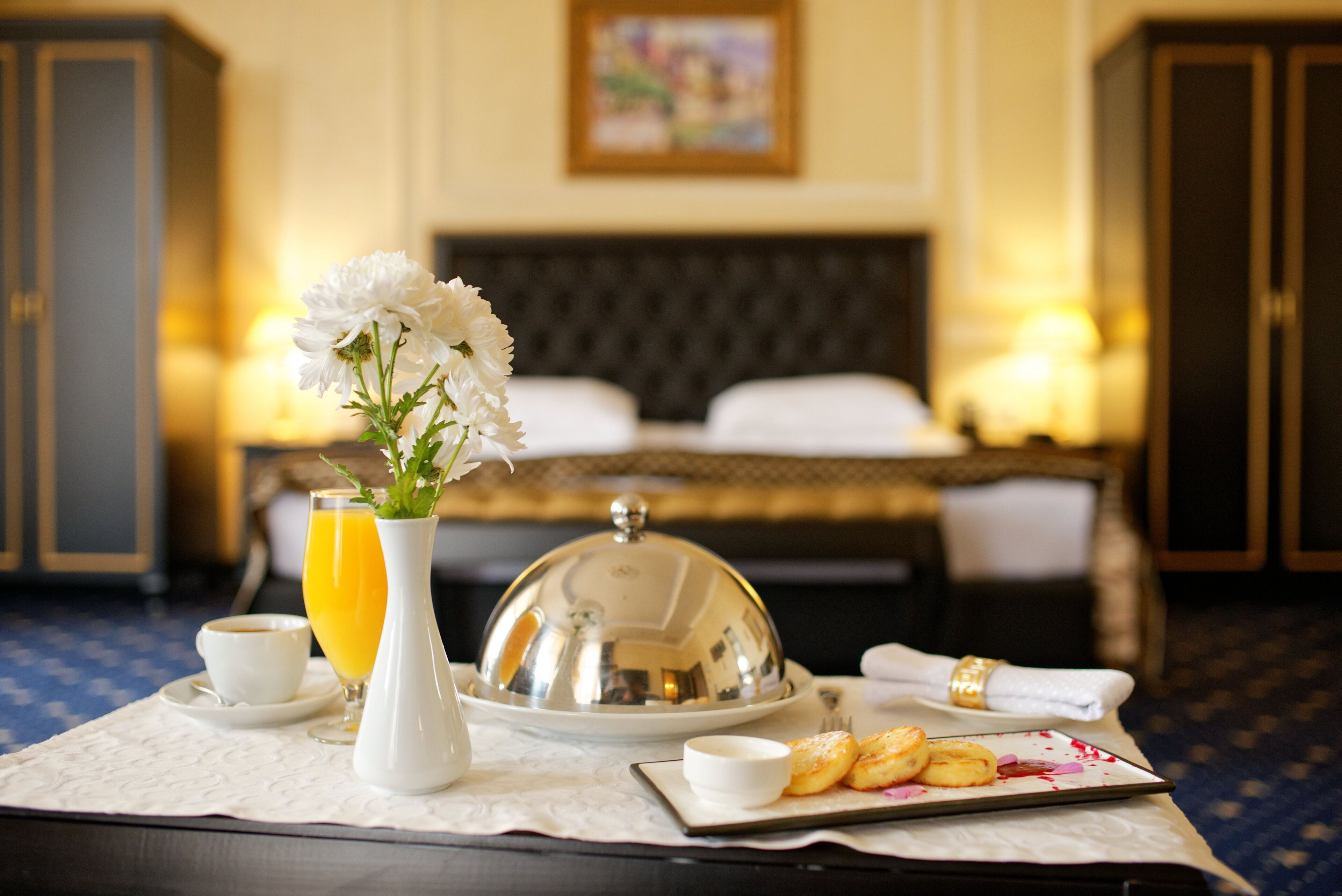 A hotel room with a neatly arranged breakfast table featuring a white vase with flowers, a glass of orange juice, a covered silver tray, a plate of pastries, and a cup of coffee.