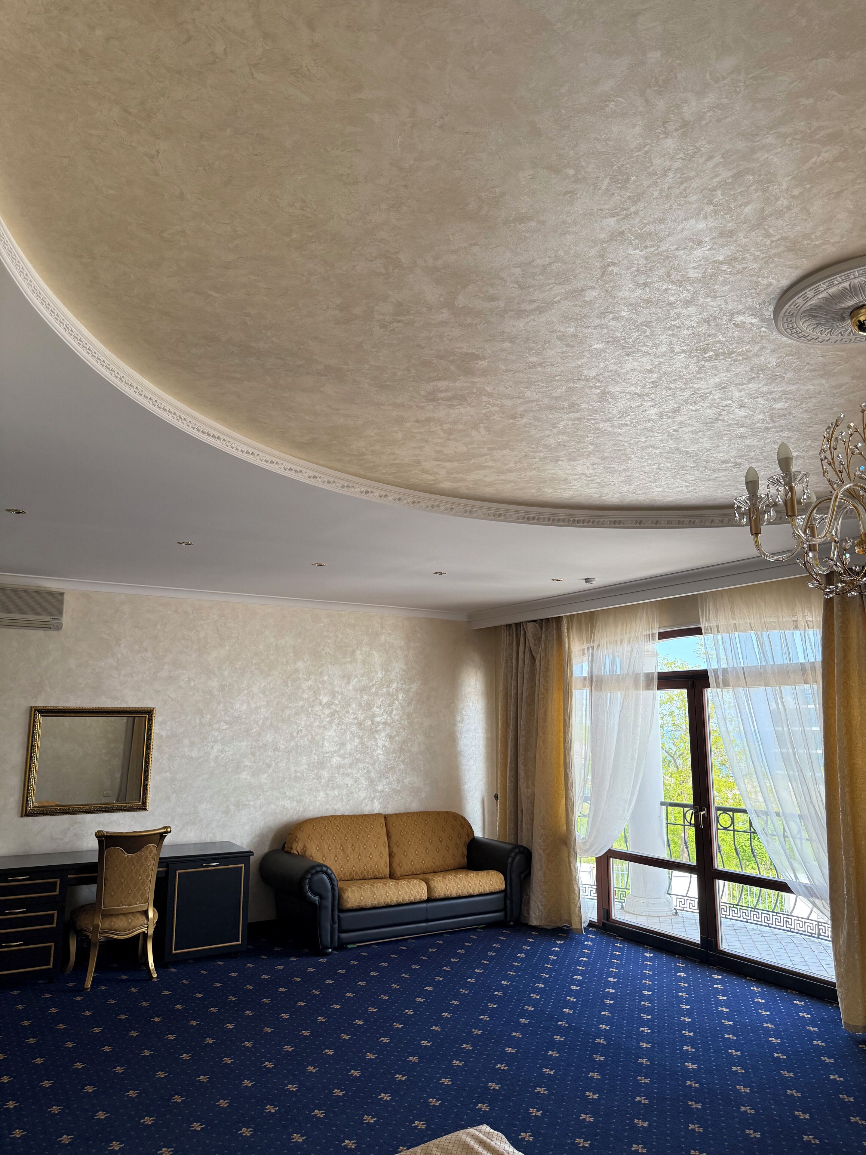 A room with a patterned carpet, a sofa, a desk with a chair, a chandelier, and a large window with curtains.