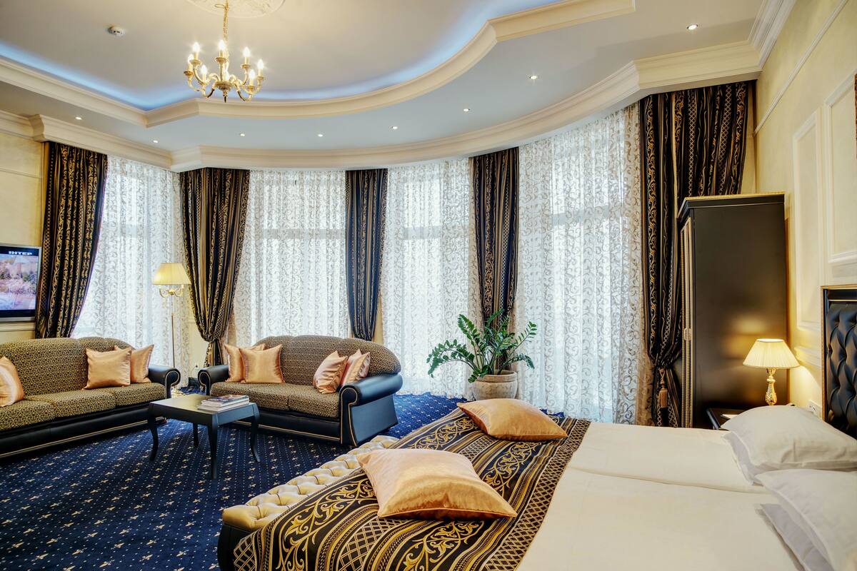 Habitación de hotel con una cama grande, un sofá, una silla y una mesita. Hay cortinas y una araña.