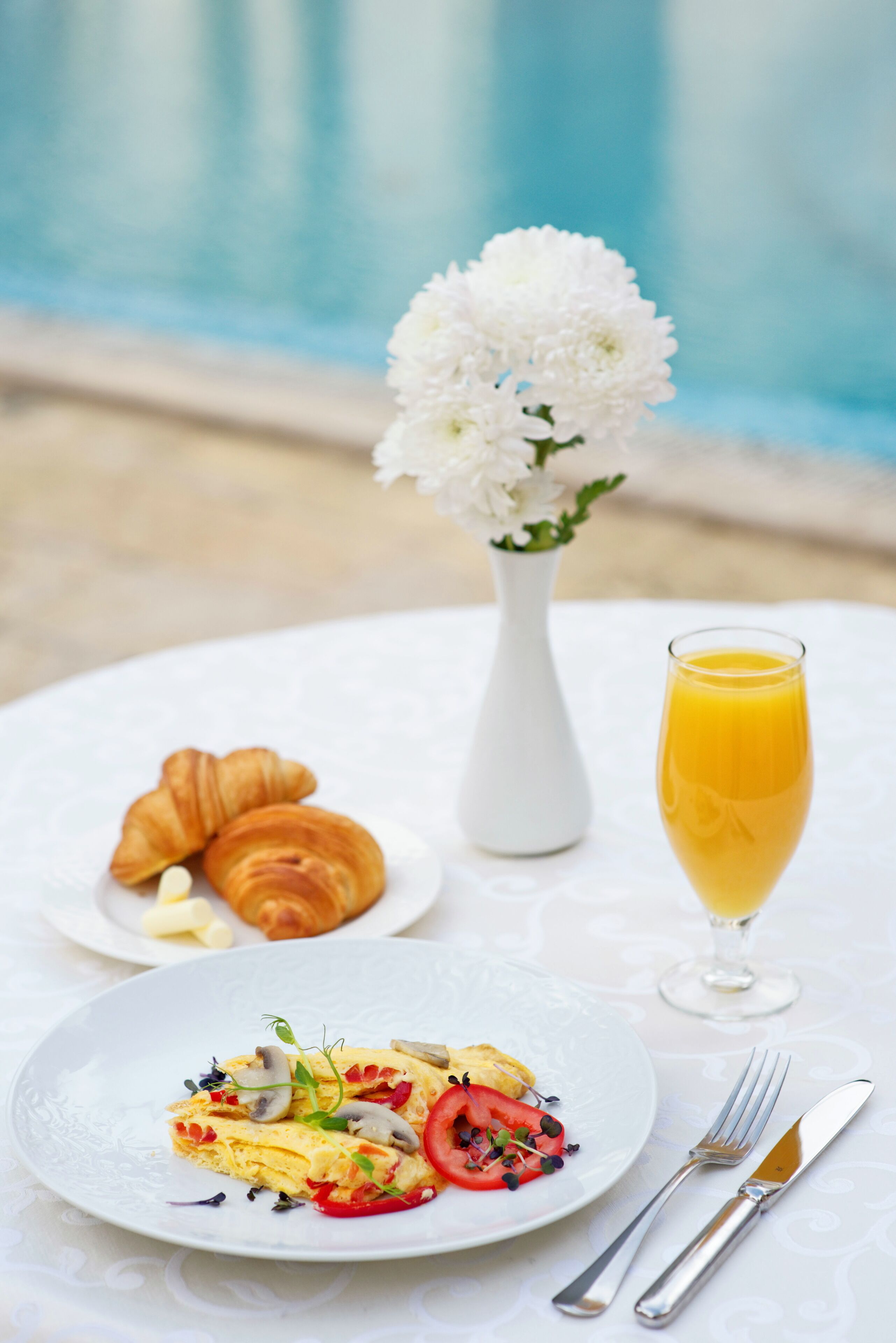 A breakfast setting with croissants, a glass of orange juice, and a plate of food by a poolside.