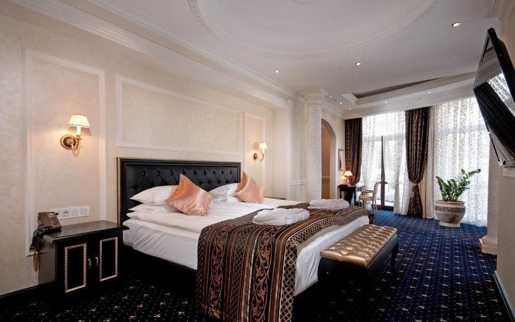 A hotel room with a large bed, a bedside table, a nightstand, a potted plant, and a large window with curtains.